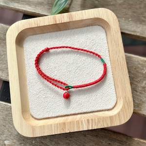 Peut inclure: Un bracelet en cordon rouge avec un petit charme de cerise rouge et vert. Le bracelet est sur un plateau en bois.