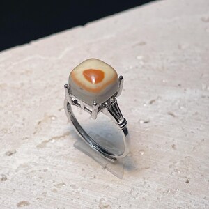 May include: A silver ring featuring a square-shaped, white and orange gemstone. The ring has a delicate design with small accents.