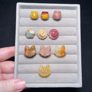 Könnte beinhalten: Eine Sammlung geschnitzter Edelsteinminiaturen in verschiedenen Formen, darunter Honigtöpfe, Blumen und Schleifen, die auf einem grauen Samttablett präsentiert werden. Die Edelsteine weisen eine Reihe von Farben auf, wie Gelb, Rosa und Orange.