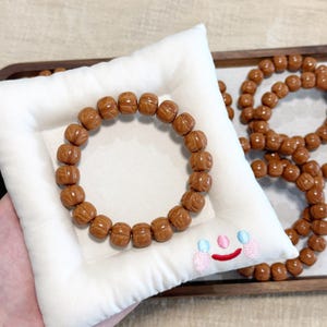 以下が含まれることがあります： 笑顔の刺繍が施された白い枕の上に、茶色のビーズブレスレットが置かれています。ブレスレットは丸く彫刻されたビーズでできています。背景には他のブレスレットもあり、ジュエリーコレクションを示唆しています。