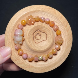 May include: A beaded bracelet with orange, pink, and white stones. The bracelet features a bear charm, a flower charm, and a gold-colored spacer with a small diamond-like accent. The bracelet is displayed on a wooden coaster.