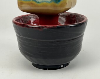 May include: A set of two ceramic bowls, one stacked on top of the other. The top bowl is a square shape with a yellow, green, and blue glaze. The bottom bowl is round with a black glaze and a red rim.