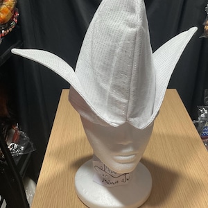 May include: A white, pointed hat with wide, wing-like flaps, displayed on a white mannequin head. The hat has a textured surface and is likely made of fabric. The mannequin is on a wooden surface.