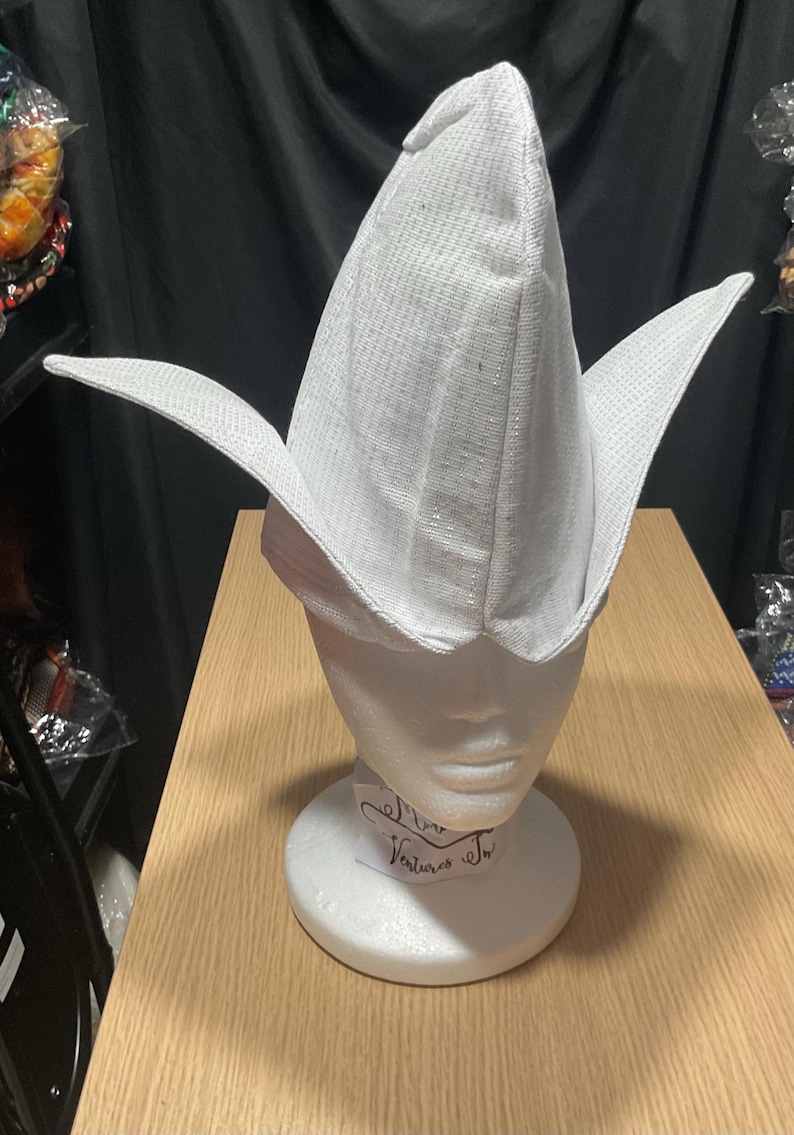 May include: A white, pointed hat with wide, wing-like flaps, displayed on a white mannequin head. The hat has a textured surface and is likely made of fabric. The mannequin is on a wooden surface.