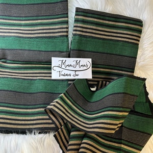 May include: Striped fabric in green, black, and beige colors. The fabric is folded and arranged on a white fluffy surface. The text "Mmau Mma's Ventures Inc" is visible on a white label.