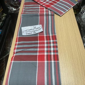 May include: A red, gray, and white plaid fabric with a matching square piece. The fabric has a rectangular shape with a repeating pattern of stripes and squares. A white label with black text is on the fabric.