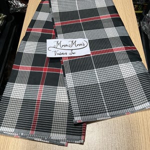 May include: A folded fabric with a black, white, and red plaid pattern. The fabric is draped over a light brown surface. A white label with the text "Mma Mma's Ventures Inc" is visible.