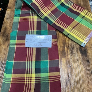 May include: Two pieces of red, green, yellow, and brown plaid fabric. The fabric is folded and laid out on a wooden surface. The text "Mama's Ventures Inc." is printed on a white label.