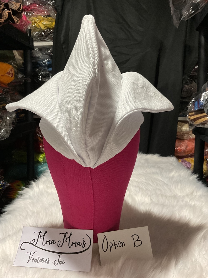 May include: A white fabric hat with a unique folded design, displayed on a pink mannequin head. The hat has a pointed top and wide, flared edges. The text "Option B" is visible on a white card. The hat is set against a backdrop of other items.