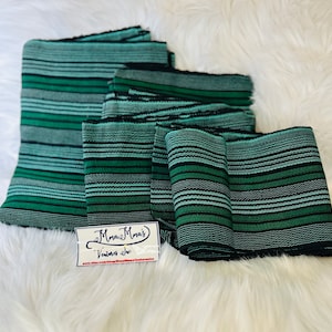 May include: A stack of five green and black striped fabric strips. The fabric is woven with a textured pattern. The strips are folded and stacked on a white fuzzy surface. A small white tag with the text "Mama Mais Ventures Inc. www.etsy.com/shop/MamaMaisVenturesInc" is in the lower left corner of the image.