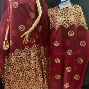 May include: A deep red fabric garment featuring intricate gold embroidery in a floral and geometric pattern along the hem and scattered circular gold embellishments across the surface. A small white tag with handwritten text is attached to the top.