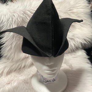 May include: A black, pointed hat with wide, curved brims, displayed on a white mannequin head. The hat has a textured surface and a subtle sparkle. The hat is likely a costume piece or a unique fashion accessory.