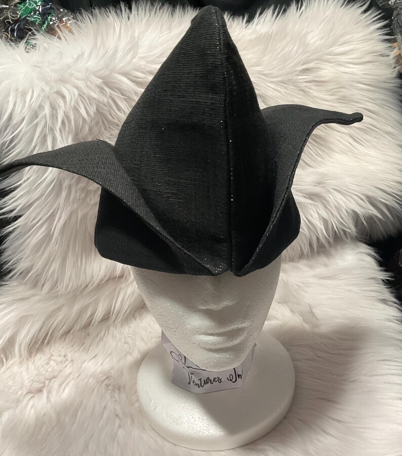 May include: A black, pointed hat with wide, curved brims, displayed on a white mannequin head. The hat has a textured surface and a subtle sparkle. The hat is likely a costume piece or a unique fashion accessory.