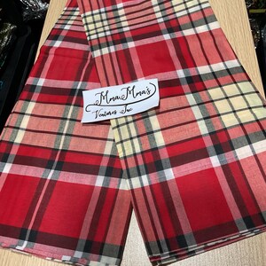 May include: Red plaid fabric with black, white, and tan accents. The fabric is folded and laid out on a wooden surface. The fabric has a repeating pattern of intersecting lines. The text "Mma Mma's Ventures Inc" is on a white label.