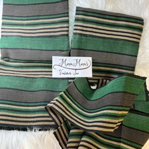 May include: Two pieces of green, black, and beige striped fabric. The fabric is folded and has a frayed edge. The fabric is labeled "Mmau Mma's Ventures Inc."