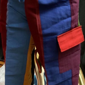 May include: Colorful patchwork pants with a drawstring waist. The pants feature panels of red, blue, purple, orange, and yellow fabric. A red pocket is visible on the side of the blue panel. The pants are displayed on a gold mannequin leg.