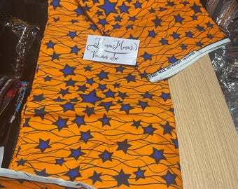 May include: Orange fabric with a repeating pattern of blue stars and wavy lines. The fabric has a white border with text. A white label with handwritten text is also visible. The fabric is folded and appears to be new.
