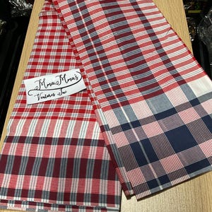 May include: Red, white, and navy blue plaid fabric. The fabric is folded and laid on a wooden surface. A white label with the text "Mman Mma's Ventures Inc" is visible.