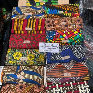 May include: A display of colorful fabric wallets with various patterns, including floral, geometric, and abstract designs. The wallets are arranged on a wooden surface, showcasing a variety of colors and styles. Some wallets have tags with letters.