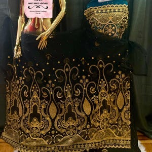 May include: A black fabric garment featuring intricate gold embroidery with swirling patterns and geometric shapes. The design includes a scalloped border at the hem and a bodice section with dense gold detailing. A label reads MMA'S MMA'S VENTURES.