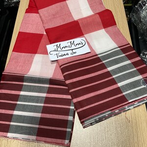 May include: A folded red, white, and gray plaid fabric. The fabric has a large check pattern in red and white, transitioning to a striped pattern in burgundy, gray, and white. A white label with black script text is visible.