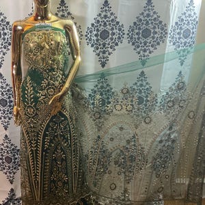 May include: A gold mannequin displays an emerald green gown with intricate gold embroidery and embellishments. The gown features a fitted bodice and a flowing skirt. A sheer, embellished fabric drapes to the side, showcasing detailed patterns. The background is a patterned curtain.
