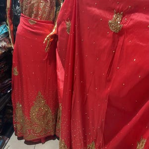 May include: A red fabric with gold embroidery and sequins. The fabric has a traditional design with a hand-shaped motif. The fabric is displayed on a mannequin.