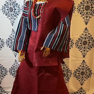 May include: A traditional maroon-colored Nigerian outfit, including a head wrap, top, skirt, and shawl. The top features striped sleeves in blue, white, and maroon. A gold necklace and bracelet are also visible. The outfit is displayed on a gold mannequin.