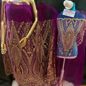 May include: A deep purple fabric featuring intricate gold embroidery and beadwork in a geometric and leaf-like pattern. The fabric is displayed on a gold-toned mannequin and a blue torso form, both featuring a white tag labeled Mma Mma's.