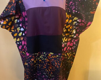May include: A long, loose-fitting dress with a V-neck and short sleeves. The dress is made of a black fabric with a colorful abstract print. The dress is layered with different colored panels, including purple, burgundy, and black.