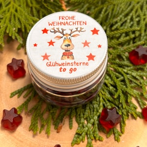 Könnte beinhalten: Ein kleines, rundes Glas mit silbernem Deckel, das eine Rentier-Illustration und den Text "Frohe Weihnachten" und "Glühweinstern to go" zeigt. Rote sternförmige Süßigkeiten sind um das Glas herum auf einem Bett aus grünem Laub verstreut.