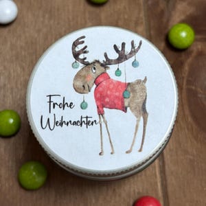 Könnte beinhalten: Ein weißer runder Behälter mit einer Illustration eines roten und braunen Elches. Der Elch trägt einen roten Pullover und hat grüne Ornamente an seinen Geweih. Der Text "Frohe Weihnachten" ist auf dem Behälter aufgedruckt.