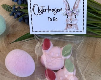 May include: An Easter-themed gift set featuring pink bunny-shaped candies with red and green accents, packaged in a clear bag. A pink egg-shaped item and a card with the text "Osterhasen To Go" and a bunny illustration are also visible.