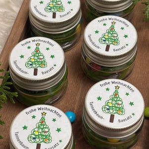 Könnte beinhalten: Fünf Glasgläser mit silbernen Deckeln, die jeweils bunte Süßigkeiten enthalten. Die Deckel sind mit einem weißen Aufkleber verziert, der einen grünen Weihnachtsbaum mit dem Text "frohe Weihnachten", "Tannenbaum" und "Bausatz" zeigt.