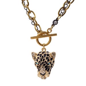 Puede incluir: Collar dorado con colgante de leopardo. El colgante es negro con detalles dorados y adornado con cristales transparentes y azules. El collar tiene un cierre de palanca y una cadena mixta dorada y negra.