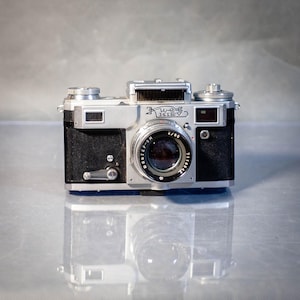 Puede incluir: Una cámara Kiev-5 vintage plateada y negra sobre una superficie reflectante. La lente tiene la inscripción "JUPITER-8M 2/50" y la palabra "KIEV" está impresa en la parte superior. Un ejemplo clásico de equipo fotográfico.
