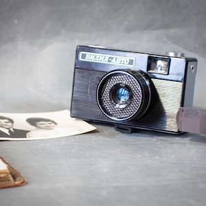 Puede incluir: Una cámara negra vintage con el texto "Вилия-Авто" se encuentra junto a un rollo de película y una fotografía en blanco y negro. La cámara tiene una lente plateada y un diseño texturizado. La película está parcialmente desenrollada.