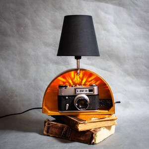 Può includere: Una lampada unica realizzata con una fotocamera vintage, con un paralume nero e una luce calda. La fotocamera è inserita in una cornice di legno, appoggiata su una pila di vecchi libri, creando un'estetica vintage.