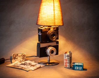 May include: A distinctive table lamp made from two vintage cameras, with a tan lampshade. The lamp has a metal base and a power cable. Old photographs and film canisters sit nearby, creating a retro aesthetic.