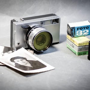 May include: A vintage silver camera with a large lens, accompanied by stacks of film boxes and black and white photographs. The film boxes are labeled "FOTO 32" and "64". A roll of film is also present.