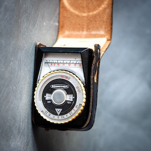 May include: A vintage black and silver Leningrad 4 light meter in a brown leather case. The meter has a circular dial with numbers and markings, and a rectangular scale at the top. The case is open, revealing the meter's face.