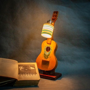Può includere: Una lampada unica, realizzata a mano, ricavata da una chitarra vintage. Il corpo della chitarra è dipinto con un motivo floreale. Il paralume è cilindrico con strisce orizzontali gialle, verdi e blu. Un libro aperto con testo è in primo piano.