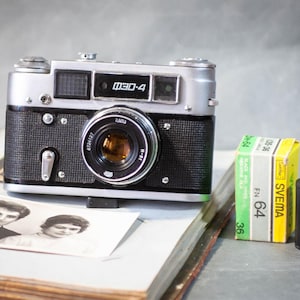 May include: Vintage silver and black camera with a lens, film rolls, and a black and white photograph. The camera has the text "Зоркий-4" on the top. The film rolls are in a yellow and green box with the text "SVEMA 64".
