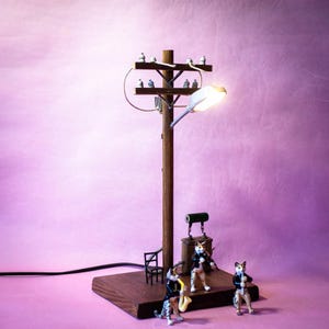 Peut inclure: Une lampe en forme de poteau électrique en bois avec un réverbère allumé, dotée de deux traverses horizontales avec de petits isolateurs et des fils. Trois figurines de chats en costume noir sont sur la base, jouant des instruments.