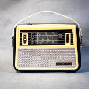 Pode incluir: Um rádio portátil vintage com uma moldura de plástico cor de creme e uma caixa preta. A parte frontal apresenta uma grelha de altifalante em metal prateado, um mostrador de sintonização de frequência com números impressos, dois botões de controlo pretos e uma pega de transporte branca. A etiqueta VEF-SPIDOLA está impressa no canto inferior direito.