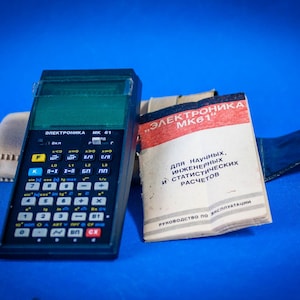 Könnte beinhalten: Ein schwarzer Elektronika MK 61 wissenschaftlicher Taschenrechner mit mehrfarbiger Tastatur, grünem Display und Netzschalter. Daneben liegen eine weiße Bedienungsanleitung mit rotem und schwarzem Text sowie eine helle Aufbewahrungstasche.