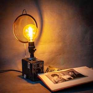 Könnte beinhalten: Eine Schreibtischlampe im Vintage-Stil, hergestellt aus einer umfunktionierten Kamera. Die Lampe hat eine große Edison-Glühbirne, einen Metallgitter-Schirm und einen schwarzen Sockel. Ein Fotoalbum mit alten Fotos liegt daneben. Die Lampe ist eingeschaltet.