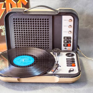 May include: A portable beige and black record player with a black vinyl record on the turntable. The device features a speaker grille, control knobs, and playback buttons. An AC/DC Highway to Hell album cover is visible in the background.