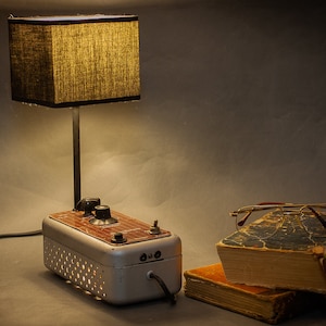 May include: A vintage-style lamp with a brown fabric shade and a metal base. The base is a repurposed metal box with a brown wood panel and a few knobs. The lamp is on a table with a stack of old books and a pair of glasses.