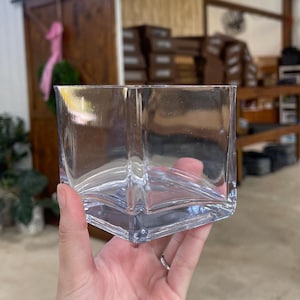 May include: A clear, square glass vase with a geometric design. The vase is held in a hand, showcasing its transparent quality. The background includes a wooden door and interior of a shop.